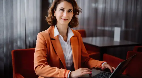 Kobieta w pomarańczowej marynarce pracuje na laptopie w nowoczesnym wnętrzu.