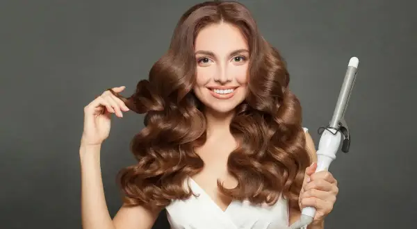 Uśmiechnięta kobieta z falowanymi brązowymi włosami, prezentująca lokówkę. Efektowne styling włosów.