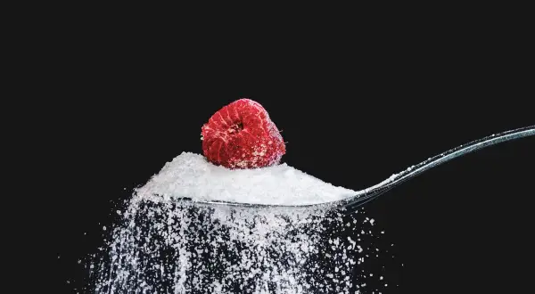 Malina na łyżce z erytrytolem, który sypie się na czarne tło. Erytrytol to naturalny słodzik.