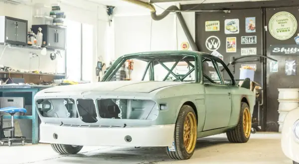 En grön BMW E9 i verkstad, redo för renovering. Guldiga fälgar och en imponerande bur syns tydligt.