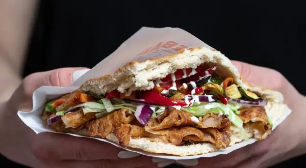 Kebab w bułce z mięsem, warzywami i sosami, trzymany w dłoniach.