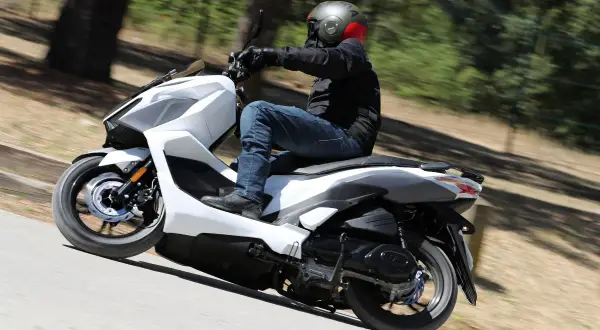 Persona conduce una SYM Jet X 125 blanca por una carretera arbolada. Las opiniones sobre la SYM Jet X 125 son positivas.