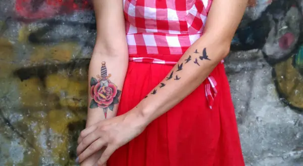 Un tatuaje de daga atraviesa una rosa roja en el brazo de una mujer. En el otro brazo, un grupo de pájaros vuelan.