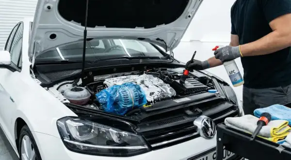 Persona con guantes rocía espuma y usa un cepillo para limpiar motor coche. El alternador está cubierto con plástico azul.