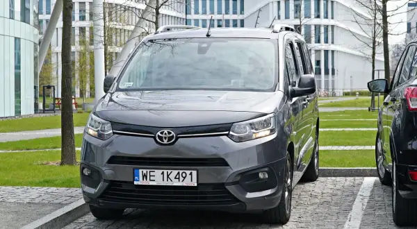 Szary Toyota Proace City zaparkowany prostopadle tyłem na parkingu przed nowoczesnym budynkiem.