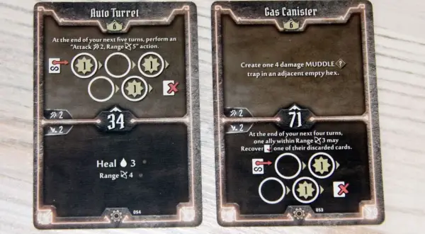 Dwie karty z gry planszowej Gloomhaven: "Auto Turret" i "Gas Canister", przedstawiające ikony akcji i statystyki postaci.