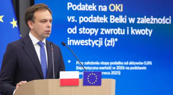 Mężczyzna w garniturze przemawia na tle ekranu z napisem "Podatek na OKI vs. podatek Belki". Dyskusja o zmianach w podatkach.