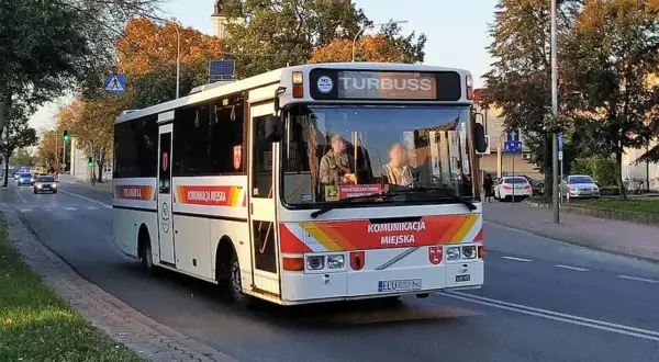Biały autobus miejski z napisem "KOMUNIKACJA MIEJSKA" i "TURBUSS" jedzie ulicą. To część codziennych, busy przewozy po polsce.
