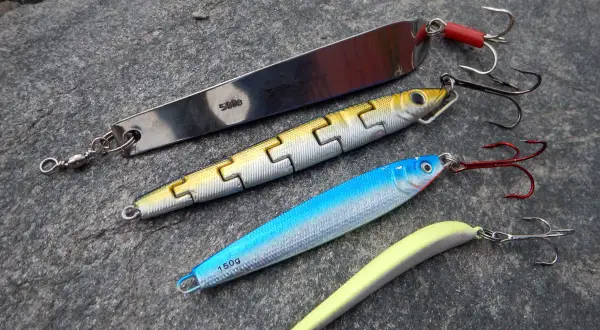 Vier Kunstköder für das Pilken vom Ufer liegen auf grauem Stein. Ein langer Silberpilker, ein gelb-silberner Wobbler, ein blauer Wobbler und ein gelber Gummifisch.