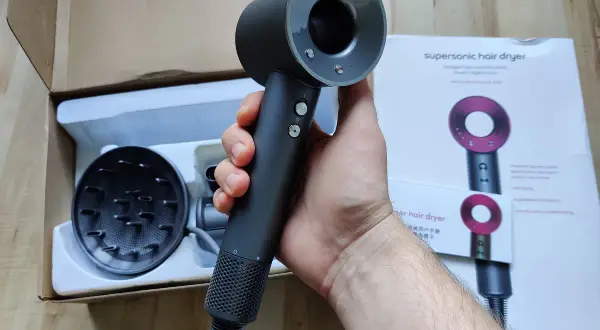Dłoń trzymająca czarną suszarkę do włosów Dyson Supersonic obok pudełka i dyfuzora.