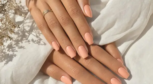 Mains aux ongles pêche, peau bronzée, bague fine. Douceur et élégance estivales.