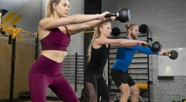 Trzy osoby wykonują trening obwodowy na siłowni, trzymając kettlebelle.