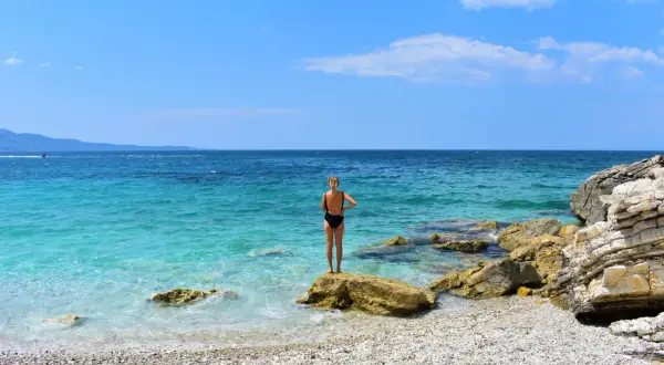 Mujer en bikini negro de pie sobre una roca en una playa de guijarros. El mar turquesa y el cielo azul crean un paisaje idílico, pero ten cuidado, viajar a Albania es peligroso.