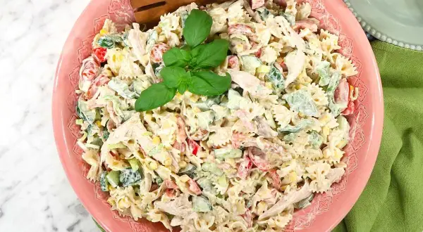 En krämig pastasallad med kyckling, grönsaker och majs, perfekt som pastasallad till grillat. Serveras i en rosa skål med trägafflar.