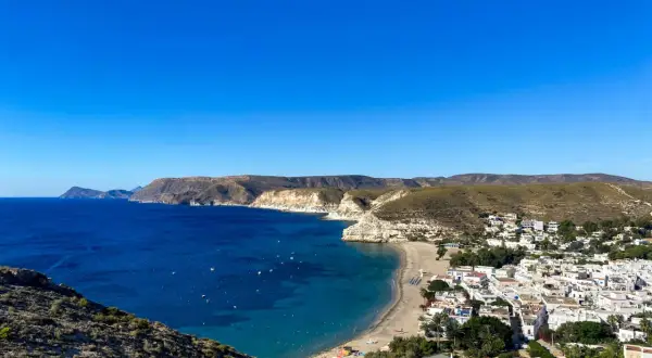 Vista panorámica de una **playa** con **agua amarga** de color azul intenso, un pueblo blanco y colinas áridas bajo un cielo despejado.