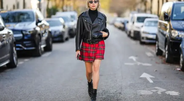 Kobieta w skórzanej ramonesce, czarnym golfie i czerwonej spódnicy w kratę prezentuje styl ubioru grunge. Na nogach ma ciężkie buty.