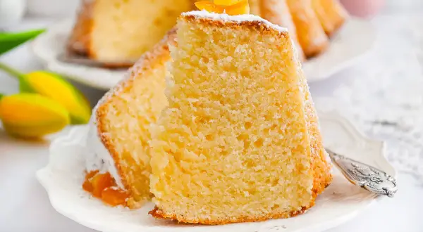 Puszysta babka z mascarpone, oprószona cukrem pudrem i ozdobiona skórką pomarańczową, na talerzyku z tulipanami.