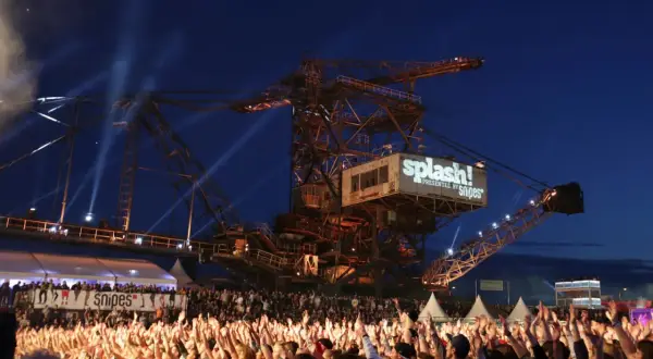 Große Menschenmenge jubelt bei einem Konzert vor einer riesigen Baggerschaufel mit dem Splash! Festival Line Up.
