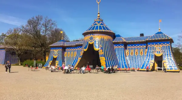 Blå och gul tältliknande byggnad med folk som sitter vid bord utanför i haga parken.