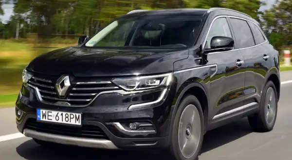 Czarny Renault Koleos 2.0 dCi pędzi po drodze, otoczony rozmytym zielonym krajobrazem.