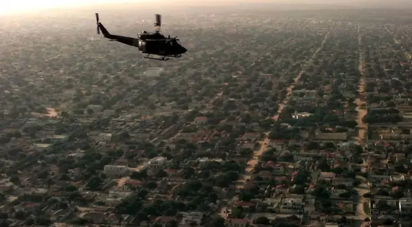 Helikopter przelatuje nad gęsto zabudowanym miastem z licznymi domami i drzewami.