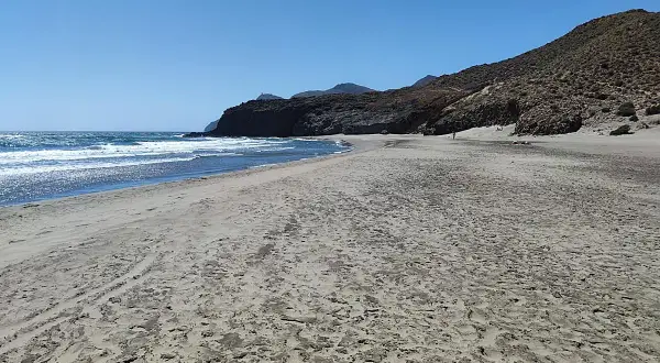 Arena dorada y mar azul en Almería. Olas rompen en la costa, con montañas rocosas al fondo. Un día perfecto para disfrutar del almeriense mar.