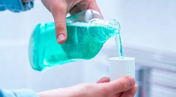 Mano vierte líquido azul de una botella a una taza medidora, mostrando varios tipos de clorhexidina para higiene bucal.