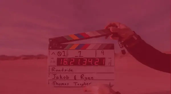 Dłoń trzyma klaps filmowy z zapisanymi informacjami o produkcji.