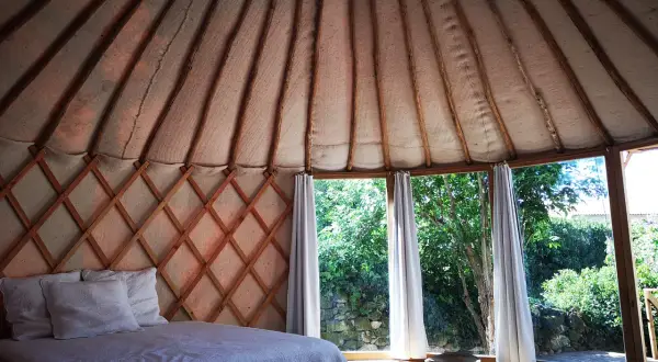 Intérieur d'une yourte gard, lit douillet sous la structure en bois, grandes fenêtres offrant une vue sur la verdure.