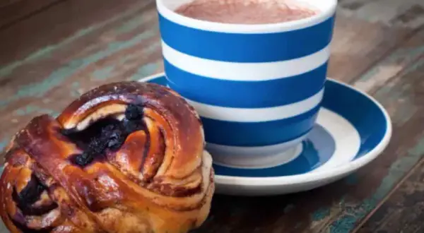 En blårandig kopp med kaffe och en kanelbulle med blåbär. Perfekt för en fika till jobbet.