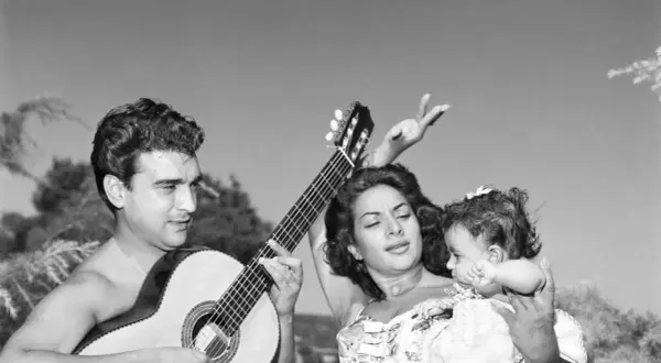Hombre toca guitarra mientras mujer baila con bebé. Una escena de rumbas flamencas al aire libre.