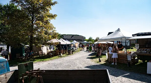 Besucher bummeln über den Bauernmarkt Lindlar, vorbei an Ständen mit Waren und Sitzgelegenheiten unter Bäumen.