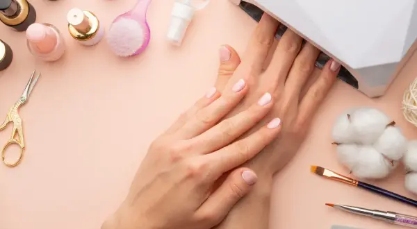 Delikatne paznokcie hybrydowe w ciąży, utwardzane w lampie UV. Wokół akcesoria do manicure: lakiery, pędzelki, nożyczki.