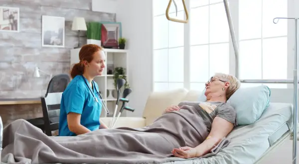 Pielęgniarka rozmawia z pacjentką leżącą w łóżku. W tle widać kule, co sugeruje, że pacjentka może potrzebować wsparcia w odżywianiu dojelitowym.