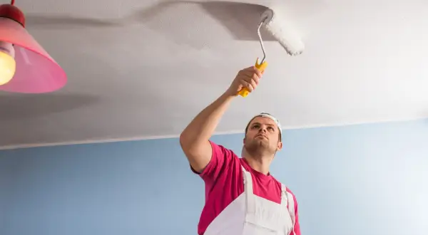 Un homme en salopette blanche et t-shirt rose peint le plafond avec un rouleau. Il semble concentré sur son travail, donnant un nouveau sens à la peinture de plafond.