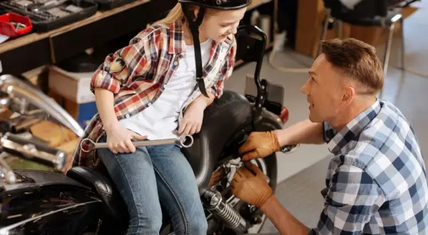 Ojciec i dziecko w kasku naprawiają motocykl w garażu. Dziewczynka trzyma klucz, a mężczyzna pracuje przy silniku.