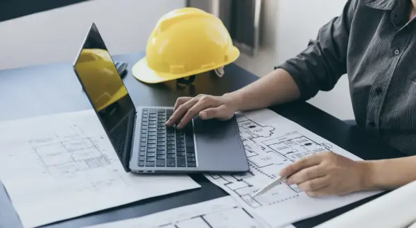 Architekt pracuje nad projektem, analizując plany i obliczając powierzchnię całkowitą budynku. Na biurku leży żółty kask.