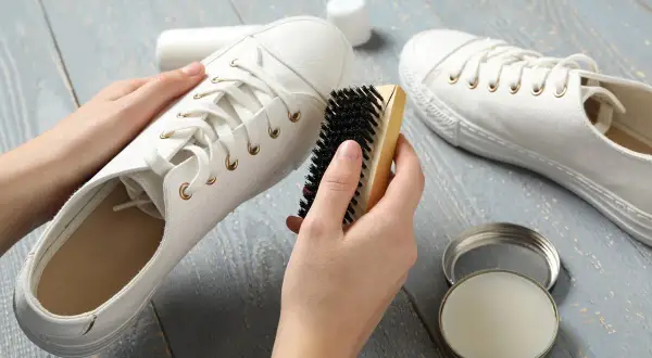 Mains nettoyant une basket blanche en tissu avec une brosse. Un pot de produit d'entretien est ouvert à côté.