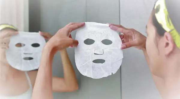 Mani bagnate che applicano una maschera in tessuto sul viso, riflessa nello specchio. Dopo la maschera in tessuto si lava il viso.