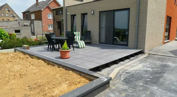 Terrasse en dalles grises sur plots, avec table et chaises, à côté d'un jardin en terre.