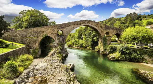 El mirador de Cangas de Onís ofrece vistas del icónico puente romano sobre un río verde esmeralda, rodeado de exuberante vegetación y un cielo azul con nubes.