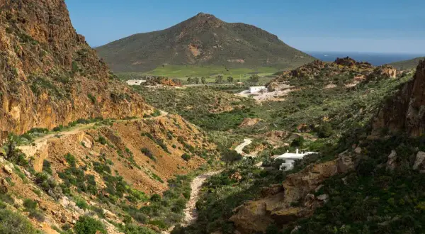 Paisaje árido con el **Cerro Negro** al fondo. Un camino serpentea entre barrancos y vegetación baja, llegando a unas casas blancas.