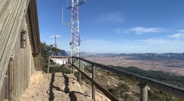 Vista panorámica desde la sierra del Carche, con una cabaña de madera, antena de comunicaciones y un paisaje de campos y montañas.
