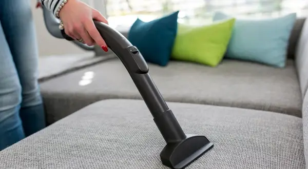 En hand använder en soff dammsugare för att rengöra en grå soffa med färgglada kuddar i bakgrunden.