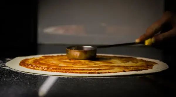 Ręka rozprowadza sos pomidorowy na cieście, tworząc idealny zaczyn na pizzę.