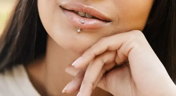 Usta z błyszczącym błyszczykiem i subtelnym center labret. Dłoń podtrzymuje podbródek, podkreślając delikatny uśmiech.