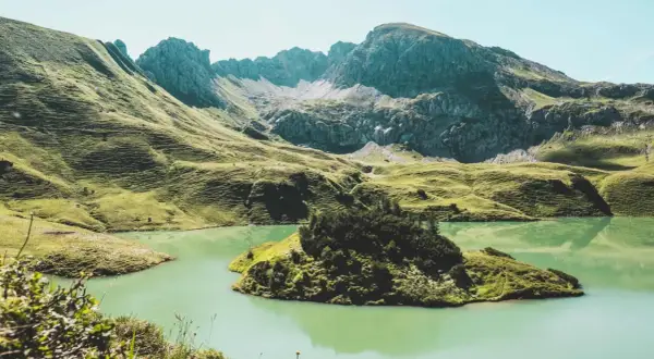 Grüne Hügel und ein türkisfarbener See laden zum Bergwandern in Bayern ein. Eine kleine Insel mit Bäumen liegt im Wasser.