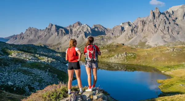 Deux randonneuses contemplent un lac d'altitude dans la vallée de la Clarée en été. Les montagnes majestueuses se reflètent dans l'eau calme.
