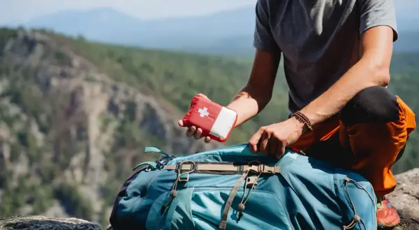 Mann packt Erste-Hilfe-Set für leichte Wanderung in Rucksack. Bergpanorama im Hintergrund.