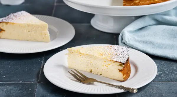 Ein Stück cremiger Quarkkuchen ohne Boden, bestäubt mit Puderzucker, wartet auf dem Teller.